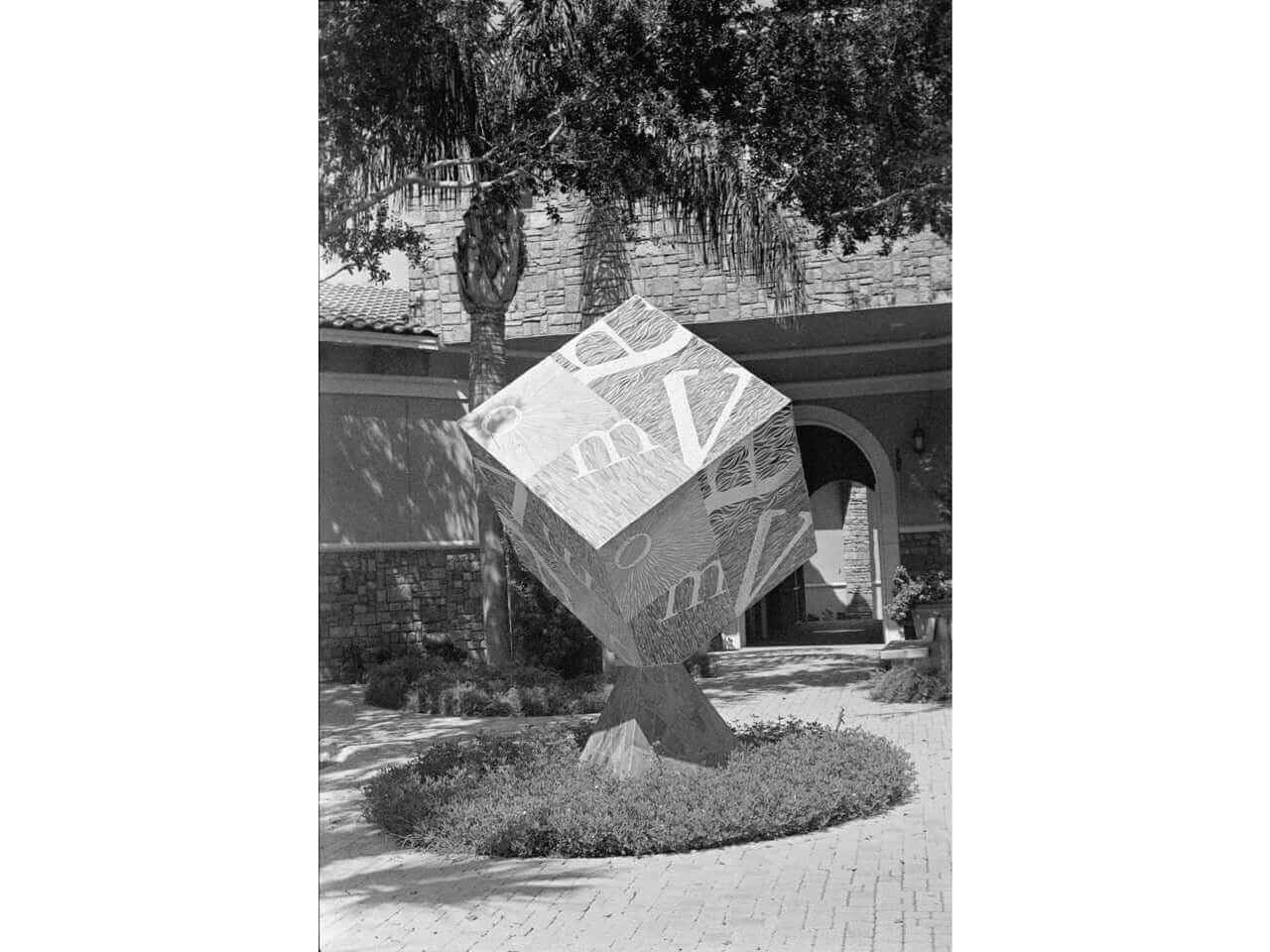 Black and white sculpture of a tilted cube with text, surrounded by greenery in a landscaped area.