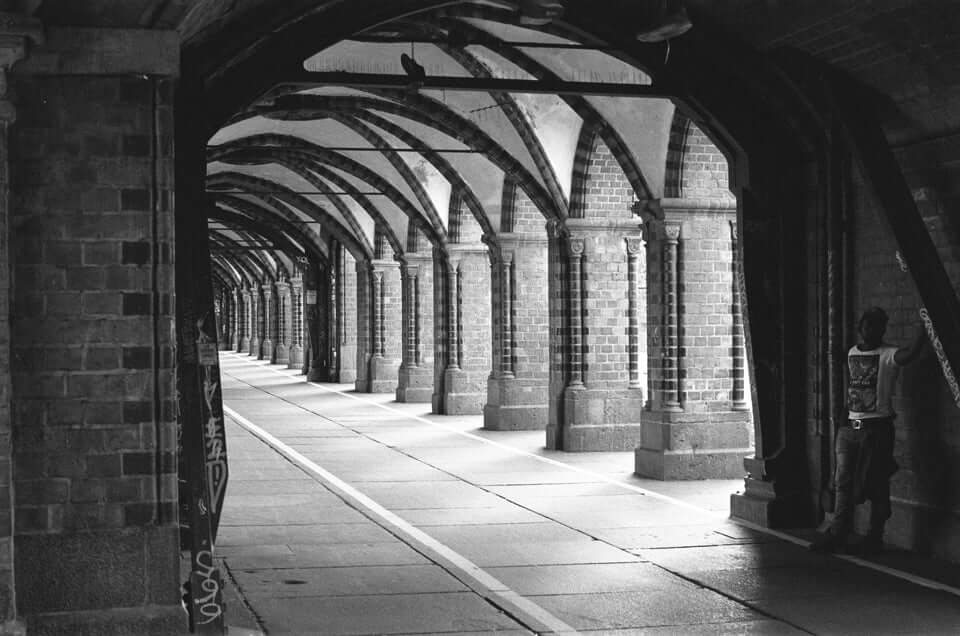 Black and white photograph of an arched brick corridor with a person leaning against the wall, showcasing urban architecture.