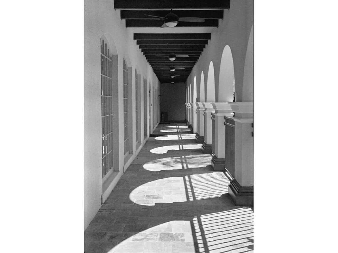 Black and white photo of a corridor with arches and shadows, showcasing architectural details and light patterns.