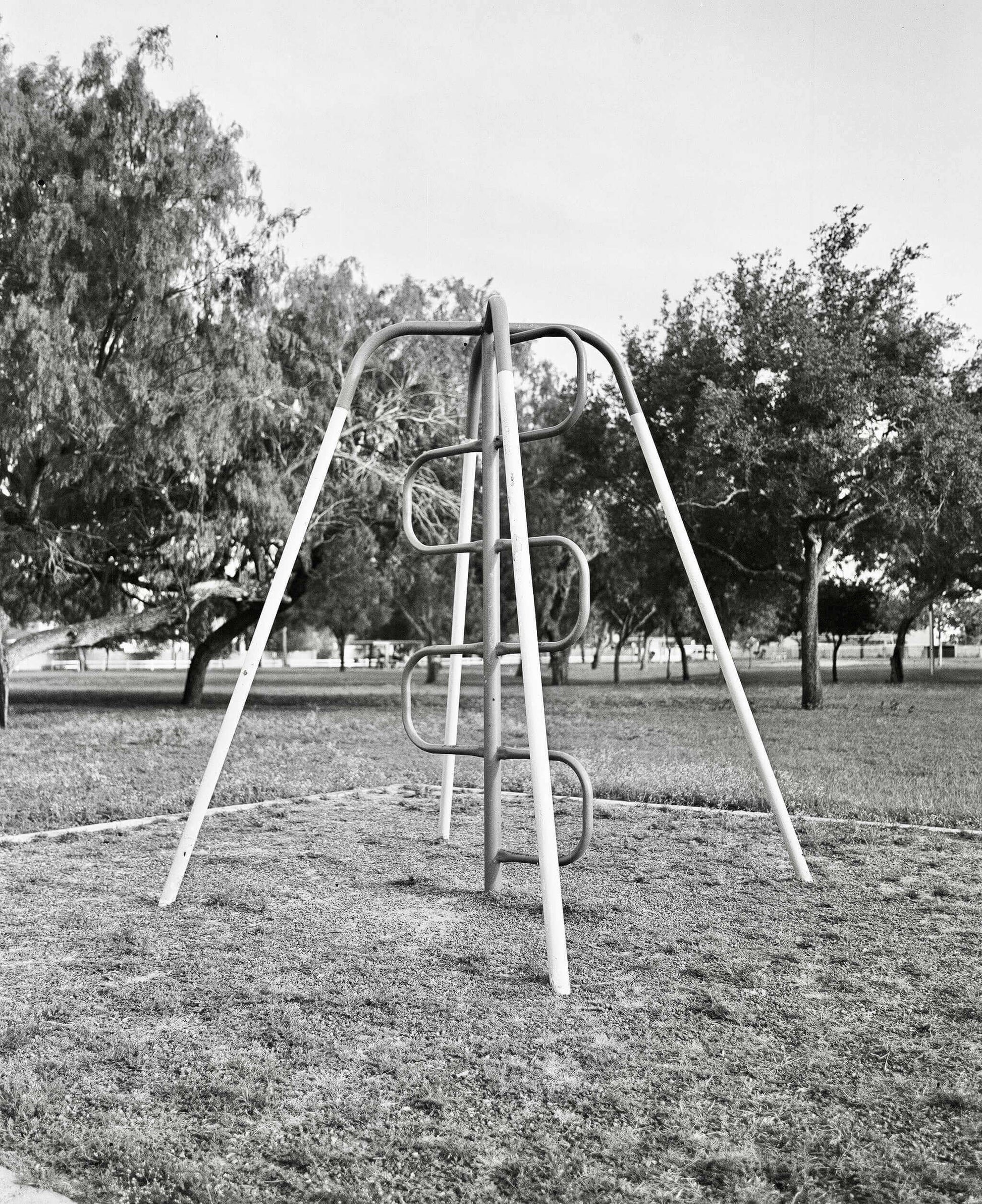 Black and white photograph of a playground swing set surrounded by trees in a grassy park.