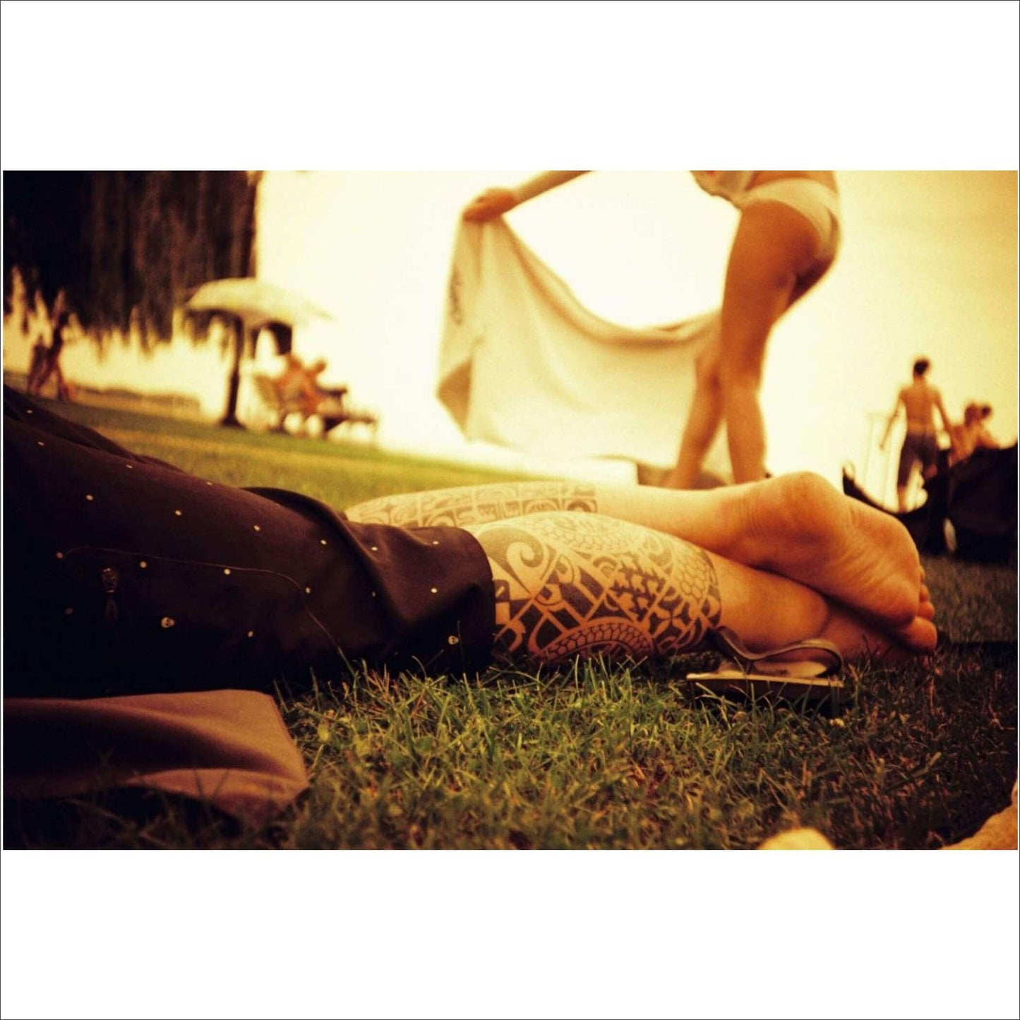 Sunlit scene featuring an arm with a tribal tattoo, relaxing on grass; a person in swimwear is in the background.